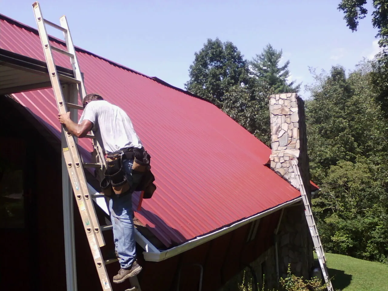 Roofer on a ladder beside a red metal roof and stone chimney — Soffit & Fascia Repair by Roofing Brothers in Quogue